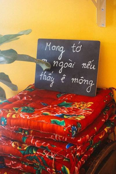 Chồng đệm ngồi m&agrave;u đỏ với hoa văn, b&ecirc;n cạnh bảng viết tay đặt tr&ecirc;n nền tường v&agrave;ng.