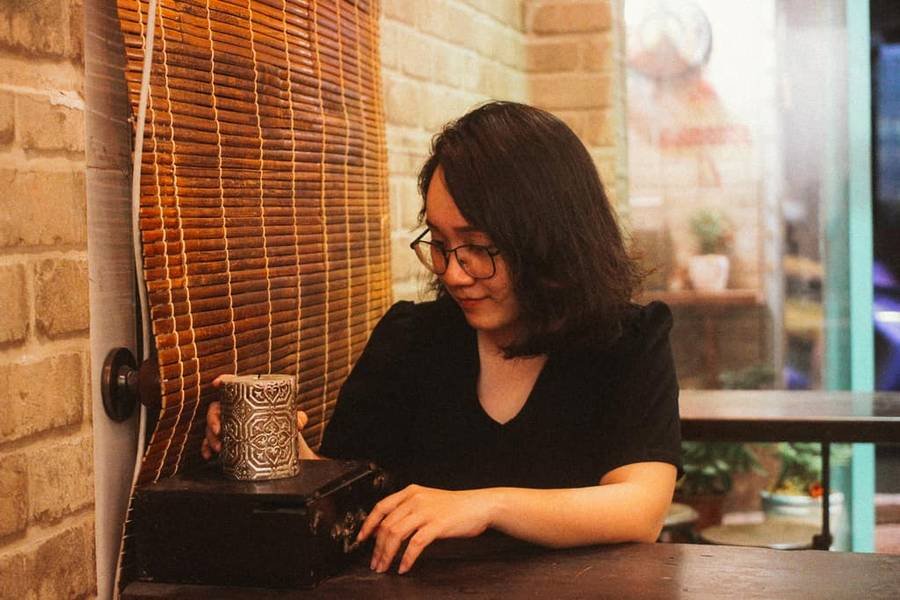 Người phụ nữ ngồi trong qu&aacute;n c&agrave; ph&ecirc;, cầm ly tr&ecirc;n b&agrave;n gỗ, b&ecirc;n cạnh r&egrave;m tre v&agrave; tường gạch.