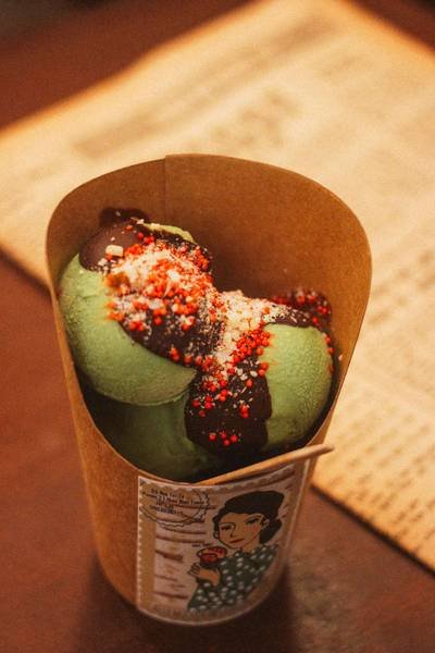 Kem m&agrave;u xanh trong cốc giấy, phủ s&ocirc; c&ocirc; la v&agrave; rắc kẹo m&agrave;u, đặt tr&ecirc;n b&agrave;n gỗ, nền l&agrave; trang giấy b&aacute;o.