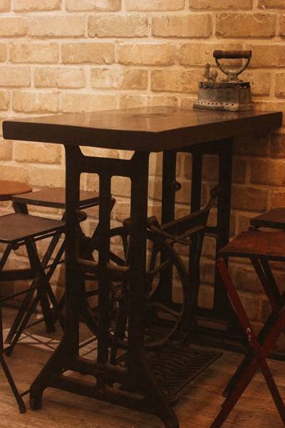 B&agrave;n l&agrave;m từ m&aacute;y may cũ, đặt trước tường gạch, tr&ecirc;n b&agrave;n c&oacute; b&agrave;n ủi cổ điển, xung quanh c&oacute; ghế gỗ.