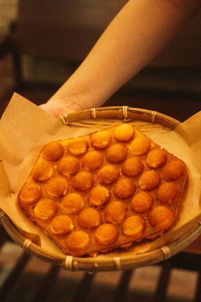 B&aacute;nh waffle tr&ograve;n nhỏ được đặt trong rổ tre, tr&ecirc;n nền giấy n&acirc;u, tay người cầm rổ.