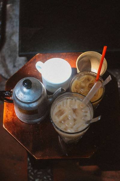 B&agrave;n gỗ với ấm tr&agrave;, cốc c&agrave; ph&ecirc; sữa đ&aacute;, cốc nước cam v&agrave; cốc sữa, &aacute;nh s&aacute;ng chiếu nhẹ từ tr&ecirc;n xuống.