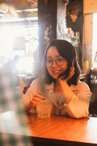 Người phụ nữ ngồi tại qu&aacute;n c&agrave; ph&ecirc;, cười v&agrave; cầm ly nước, &aacute;nh s&aacute;ng ấm &aacute;p chiếu v&agrave;o kh&ocirc;ng gian xung quanh.