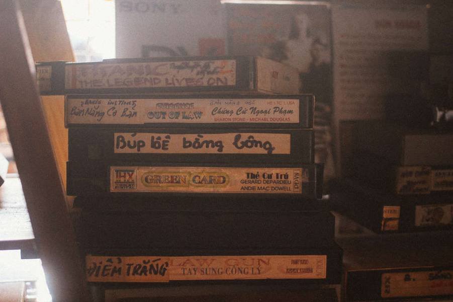 Một chồng băng VHS cũ với nhiều tựa phim kh&aacute;c nhau, được xếp chồng l&ecirc;n nhau tr&ecirc;n b&agrave;n gỗ trong &aacute;nh s&aacute;ng mờ.