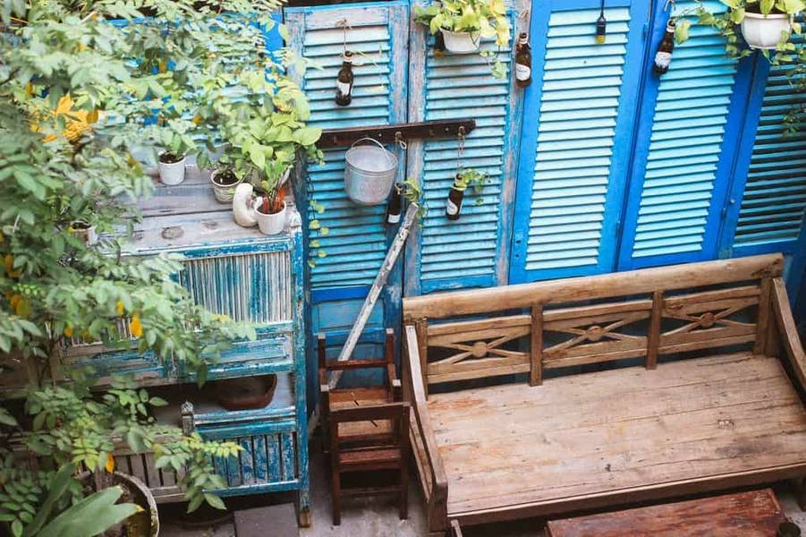Khu vườn nhỏ với ghế gỗ, cửa chớp xanh, chậu c&acirc;y treo v&agrave; c&acirc;y xanh xung quanh.