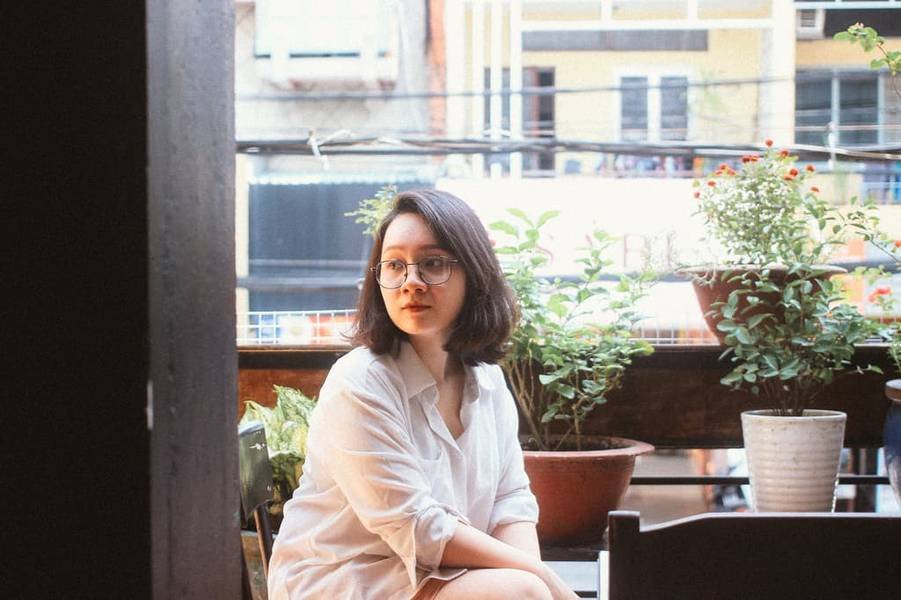 Người ngồi tr&ecirc;n ban c&ocirc;ng với &aacute;o sơ mi trắng, đằng sau l&agrave; c&aacute;c chậu c&acirc;y v&agrave; t&ograve;a nh&agrave;.