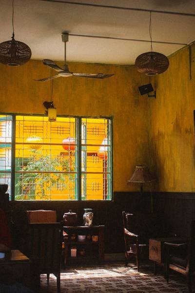 Căn ph&ograve;ng với tường v&agrave;ng, cửa sổ k&iacute;nh, b&agrave;n ghế gỗ, đ&egrave;n treo v&agrave; quạt trần, &aacute;nh s&aacute;ng tự nhi&ecirc;n từ cửa sổ.