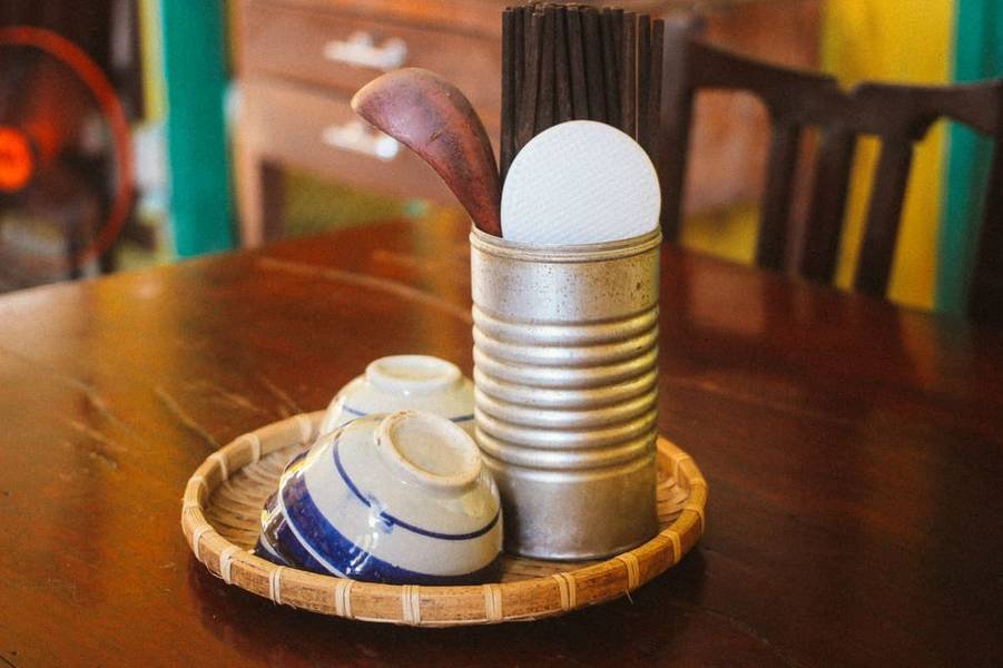 Đồ d&ugrave;ng nh&agrave; bếp tr&ecirc;n khay tre: b&aacute;t sứ, hộp đựng đũa, th&igrave;a gỗ v&agrave; đĩa nhựa đặt tr&ecirc;n b&agrave;n gỗ.