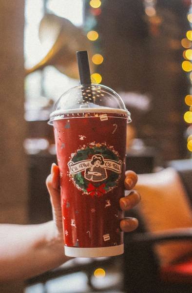 Người cầm ly nhựa m&agrave;u đỏ c&oacute; nắp đậy v&agrave; ống h&uacute;t, nền c&oacute; đ&egrave;n v&agrave; nội thất mờ mờ.