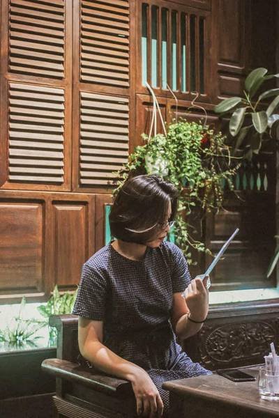 Người ngồi trong qu&aacute;n c&agrave; ph&ecirc; cổ, cầm vật dụng, nền l&agrave; cửa gỗ v&agrave; c&acirc;y xanh trang tr&iacute;.