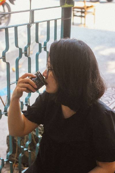 Người ngồi uống nước từ cốc nhỏ cạnh lan can, mặc &aacute;o đen, t&oacute;c ngắn, ngồi b&ecirc;n ngo&agrave;i v&agrave;o ban ng&agrave;y.