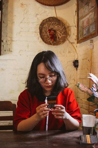 Người mặc &aacute;o đỏ ngồi trong qu&aacute;n c&agrave; ph&ecirc;, nh&igrave;n v&agrave;o một tấm thẻ tr&ecirc;n tay, xung quanh c&oacute; c&acirc;y cảnh v&agrave; đồ trang tr&iacute;.