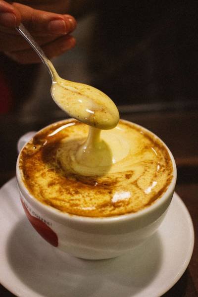 T&aacute;ch c&agrave; ph&ecirc; trứng với lớp bọt kem v&agrave;ng, một chiếc th&igrave;a đang khuấy ph&iacute;a tr&ecirc;n, đặt tr&ecirc;n đĩa trắng.