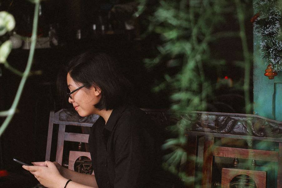 Người ngồi tr&ecirc;n ghế cầm điện thoại, ph&iacute;a trước c&oacute; c&acirc;y xanh v&agrave; đồ trang tr&iacute;.