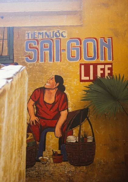 Tranh tường vẽ người phụ nữ ngồi cạnh giỏ đồ, nền chữ "Tiệm nước Sai-Gon Life" tr&ecirc;n tường m&agrave;u v&agrave;ng.