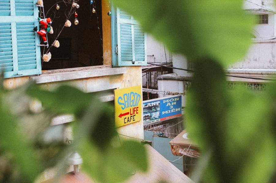 Cửa sổ xanh với bảng hiệu "Saigon Life Cafe", ph&iacute;a trước c&oacute; l&aacute; c&acirc;y, d&acirc;y đ&egrave;n trang tr&iacute; treo b&ecirc;n tr&ecirc;n.