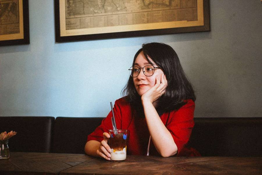 Người phụ nữ mặc &aacute;o đỏ, đeo k&iacute;nh, ngồi trong qu&aacute;n c&agrave; ph&ecirc; với ly đồ uống đặt tr&ecirc;n b&agrave;n gỗ.