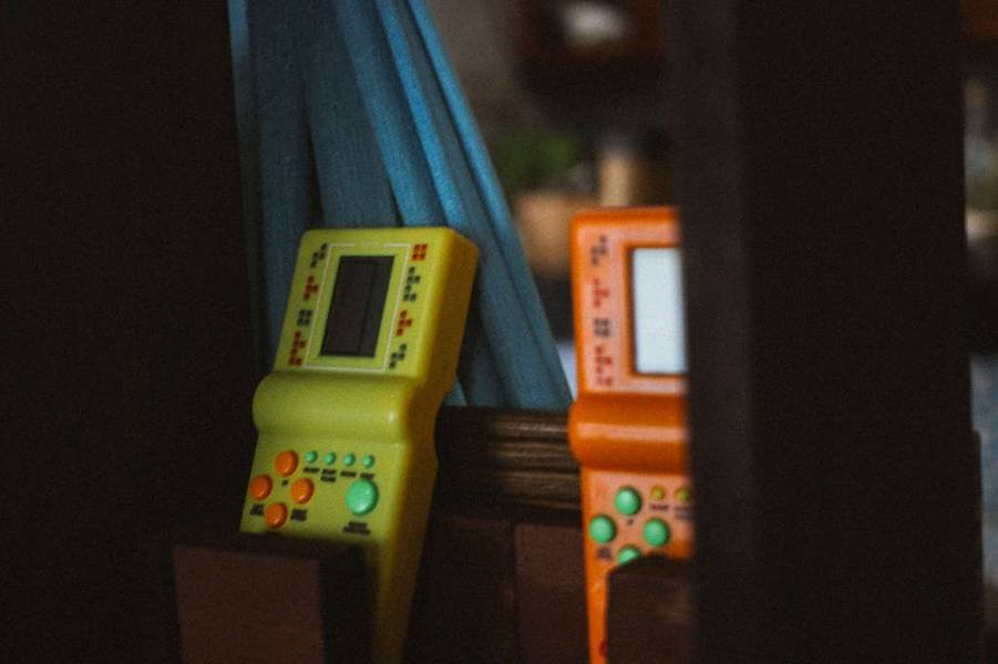 Hai m&aacute;y chơi game cầm tay m&agrave;u v&agrave;ng v&agrave; cam đặt cạnh nhau tr&ecirc;n một bề mặt gỗ.