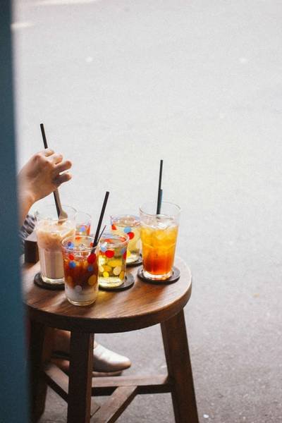 Năm ly tr&agrave; sữa đầy m&agrave;u sắc với ống h&uacute;t tr&ecirc;n b&agrave;n gỗ tr&ograve;n, tay người đang khuấy một ly.