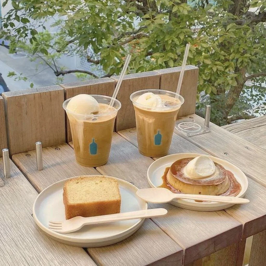 Hai ly c&agrave; ph&ecirc; đ&aacute; v&agrave; b&aacute;nh ngọt tr&ecirc;n b&agrave;n gỗ ngo&agrave;i trời, c&oacute; c&acirc;y xanh ph&iacute;a sau.