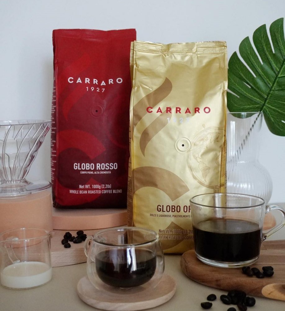 1-kg-ca-phe-pha-may-carraro Hai gói cà phê Carraro cạnh ly cà phê và lá cây, bối cảnh trang trí hiện đại.