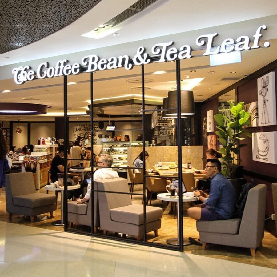 Top 32 thương hiệu nhượng quyền cafe siêu lợi nhuận 16 Quán cà phê với khách ngồi thư giãn bên trong, có bảng hiệu lớn phía trên và cây xanh trang trí.
