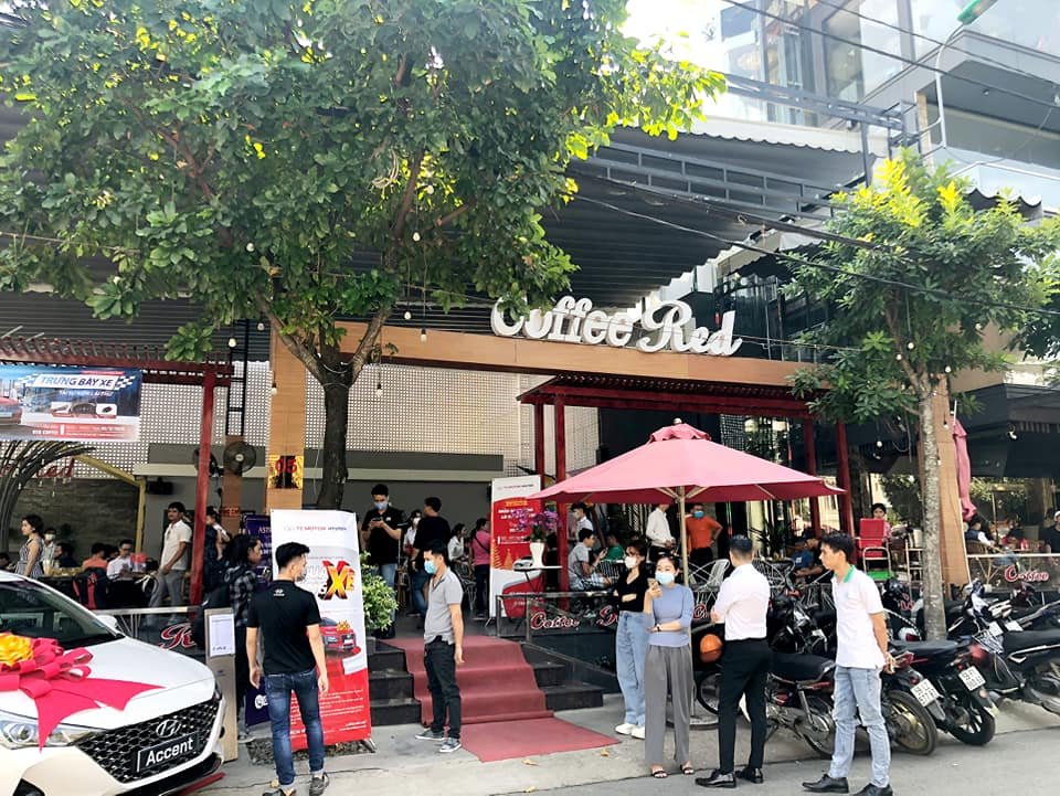 Quán cà phê đông đúc với nhiều người đứng và ngồi, bên ngoài có xe hơi trưng bày và ô che nắng màu đỏ.