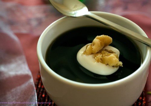 Cốc đựng món tráng miệng màu đen với lớp kem trắng và hạt óc chó trên bề mặt, kèm một chiếc thìa kim loại.