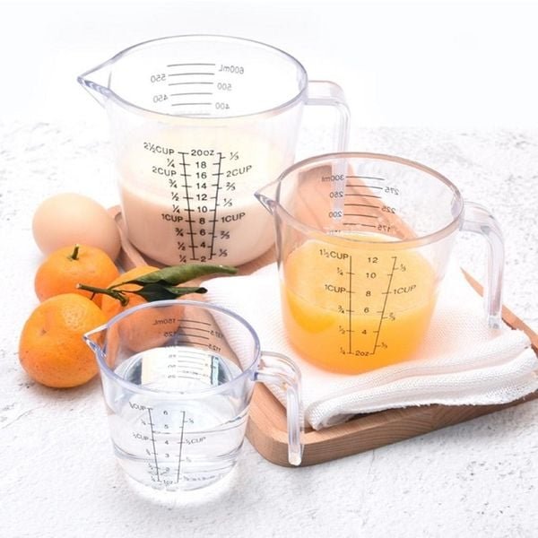 Ba ly đo lường chứa sữa, nước cam và nước, đặt trên khăn và thớt, cạnh quả cam và trứng.