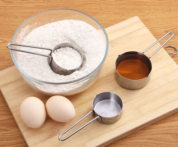 Bát bột mì, hai quả trứng, và hai cốc đo chứa chất lỏng trên bàn gỗ, chuẩn bị cho nấu ăn.