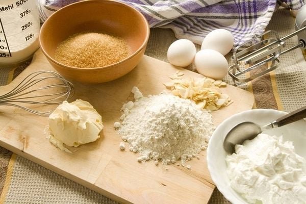Nguyên liệu làm bánh gồm bột mì, đường, trứng, bơ, kem tươi, hạnh nhân lát và các dụng cụ nhà bếp trên bàn.