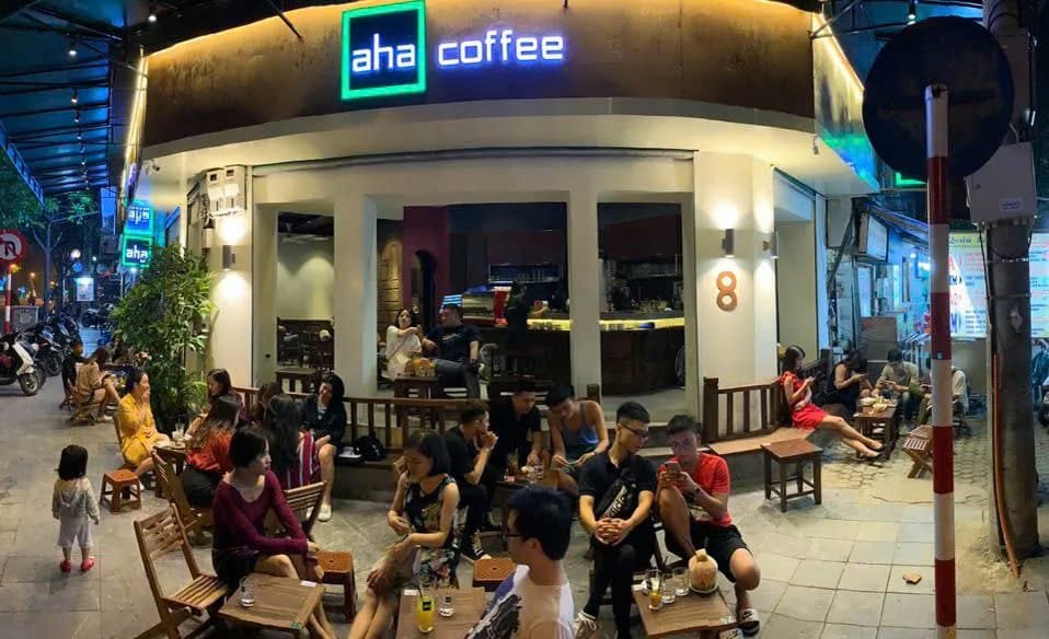 Quán cà phê đông khách ngồi ngoài trời buổi tối, ánh sáng ấm áp, biển hiệu "aha coffee" phát sáng.