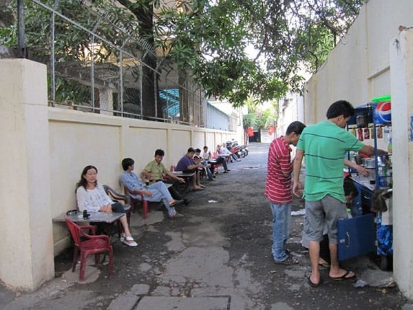 Người dân ngồi uống cà phê ở một quán vỉa hè trong con hẻm nhỏ với cây cối xung quanh.