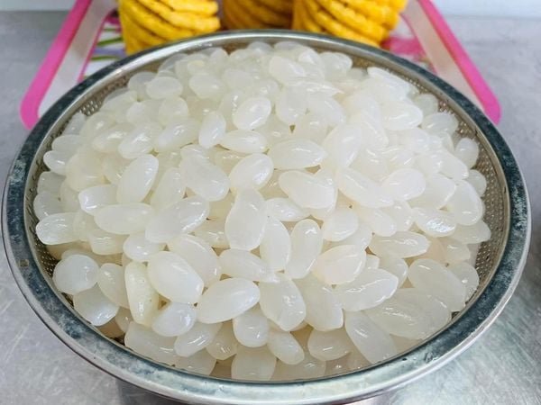 Rổ đựng những hạt màu trắng bóng, có hình dạng giống hạt bắp, đặt trên bàn với một số đồ vật phía sau.