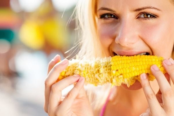 Người phụ nữ đang ăn bắp ngô, cầm trên tay, bối cảnh ngoài trời sáng sủa.
