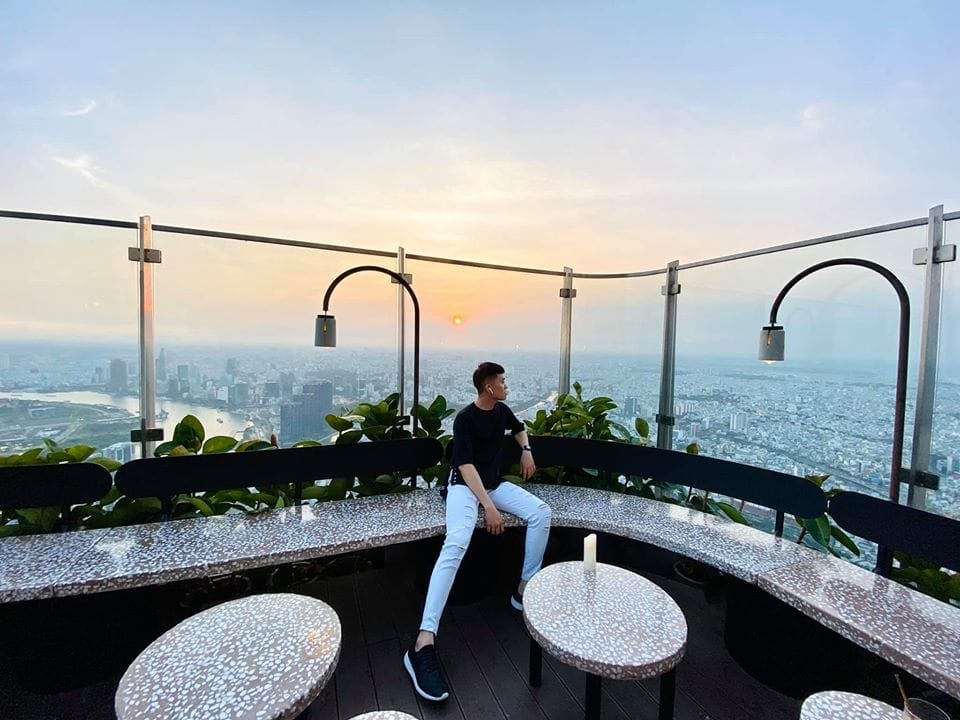 Người ngồi trên sân thượng quán cà phê, nhìn ra cảnh hoàng hôn trên thành phố với hàng rào kính bao quanh.