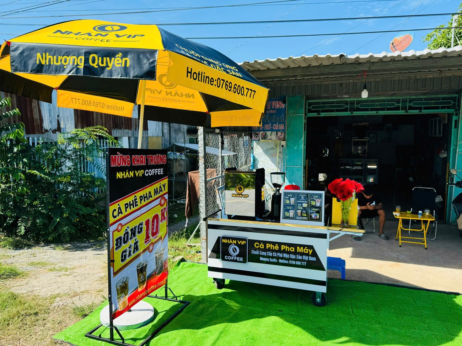 Quầy cà phê lưu động với ô vàng lớn, bảng hiệu khai trương, và hoa trang trí trước cửa hàng nhỏ.
