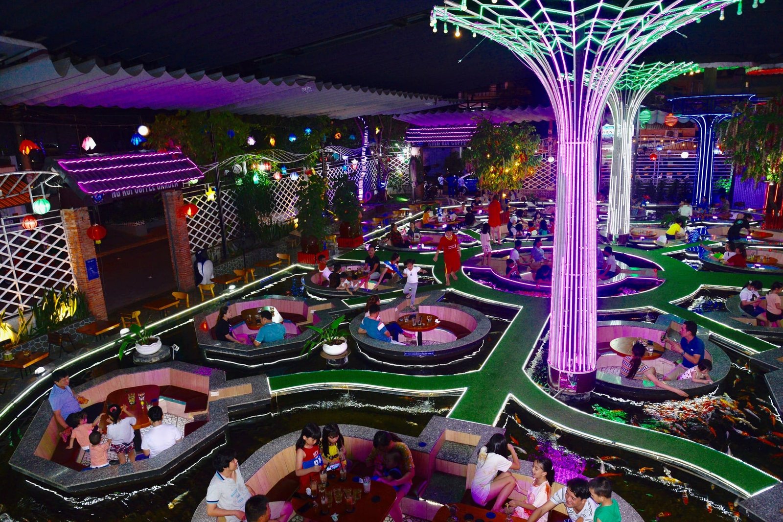 Quán cà phê ngoài trời với đèn neon sáng, nhiều bàn tròn cạnh hồ cá, khách hàng ngồi trò chuyện vui vẻ.