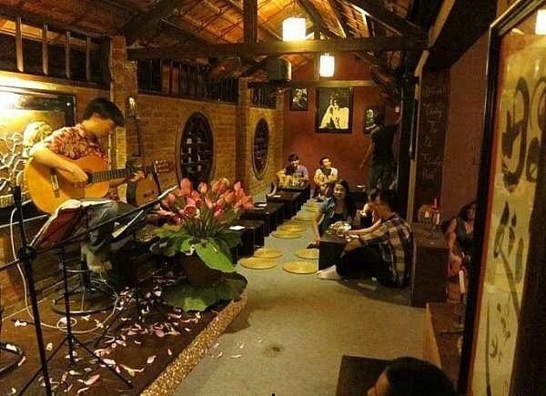 Một người chơi guitar trên sân khấu trong quán cà phê ấm cúng, với khán giả ngồi thưởng thức âm nhạc.