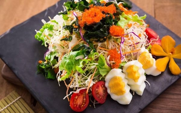 Đĩa salad rau củ tươi với trứng cắt l&aacute;t v&agrave; trứng c&aacute;, trang tr&iacute; với hoa v&agrave; c&agrave; chua tr&ecirc;n b&agrave;n gỗ.