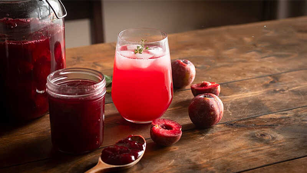 Ly nước ép mận đỏ và lọ mứt trên bàn gỗ, xung quanh là những quả mận tươi.