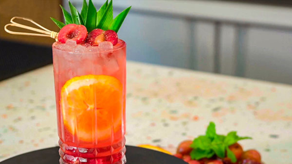 Ly cocktail màu hồng với lát cam, trang trí quả mọng và lá xanh, đặt trên bàn đá hoa văn.