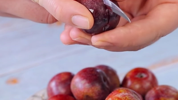 Người cắt trái mận đỏ bằng dao nhỏ, bên dưới có một đĩa trái mận khác.