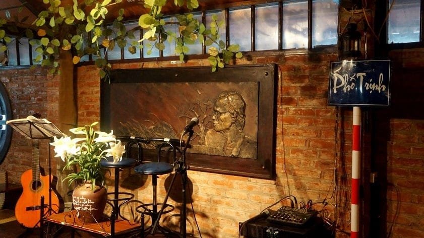 Góc quán cà phê với cây đàn guitar, bàn ghế, bức phù điêu trên tường và giàn cây leo trang trí.