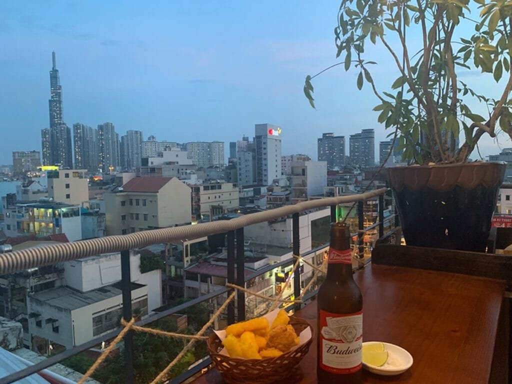 Bữa ăn nhẹ trên ban công với ly bia, nhìn ra thành phố với các tòa nhà cao tầng và bầu trời trong xanh.