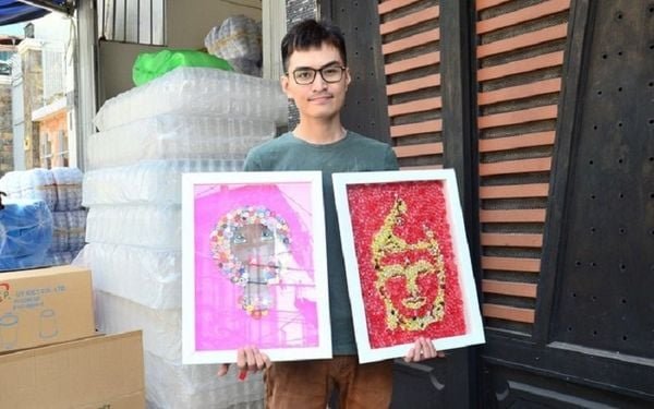 Người cầm hai bức tranh khảm màu sắc rực rỡ, đứng trước cửa gỗ và các chai nhựa xếp chồng.