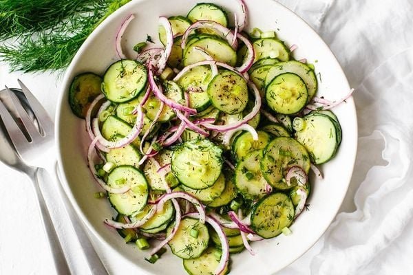 Đĩa salad gồm dưa leo thái lát, hành tây đỏ và thì là, bày biện trên nền vải trắng với dụng cụ ăn bên cạnh.