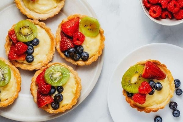 Đĩa b&aacute;nh tart tr&aacute;i c&acirc;y với d&acirc;u, kiwi, việt quất, b&ecirc;n cạnh l&agrave; ch&eacute;n m&acirc;m x&ocirc;i đỏ.