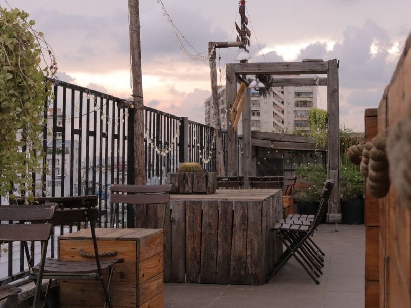 Quán cà phê ngoài trời trên tầng thượng với bàn ghế gỗ, cây xanh, và dây đèn trang trí, dưới bầu trời hoàng hôn.