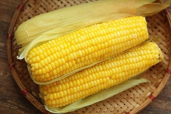 Hai bắp ngô vàng tươi đặt trên rổ tre, một bắp còn nguyên vỏ, trên nền gỗ nâu.